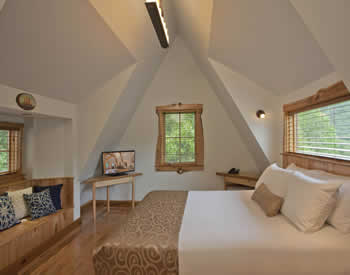 Cozy room with light blue walls and steep sloped roof. One queen bed and build-in bench with many soft pillows.