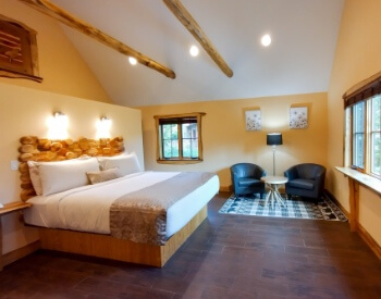 a bedroom with a bed, lamps above the bed, 2 armchairs and a small table in the corner, windows along one wall, and wood beams on the ceiling.