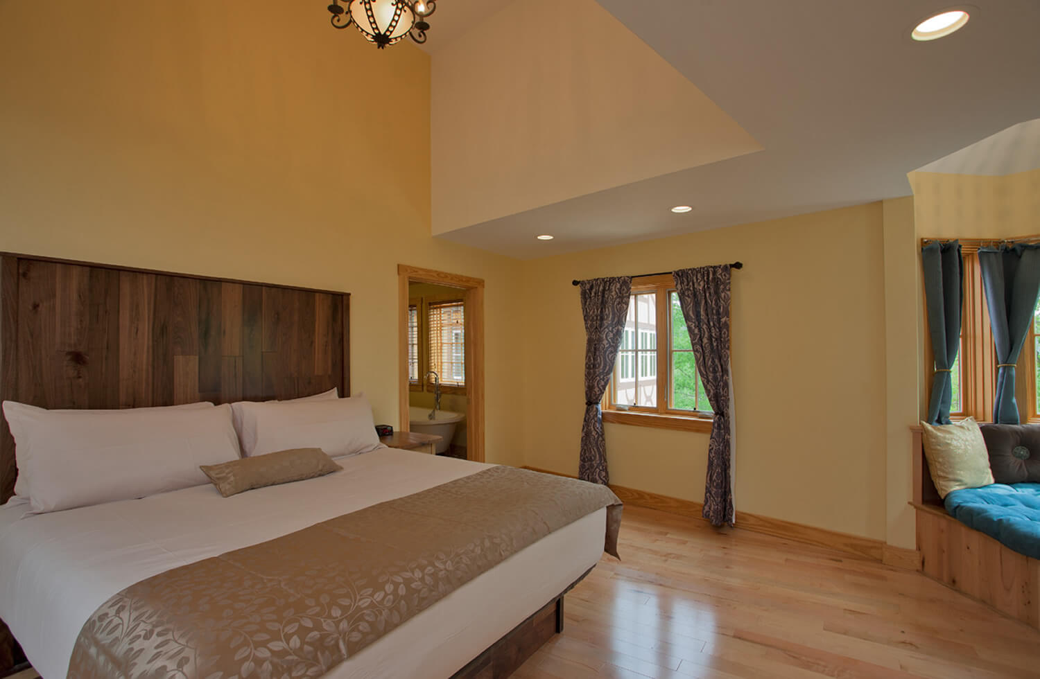 A yellow guestroom has white accented on unique ceiling; a huge well-made white bed with tan accent pillow and throw