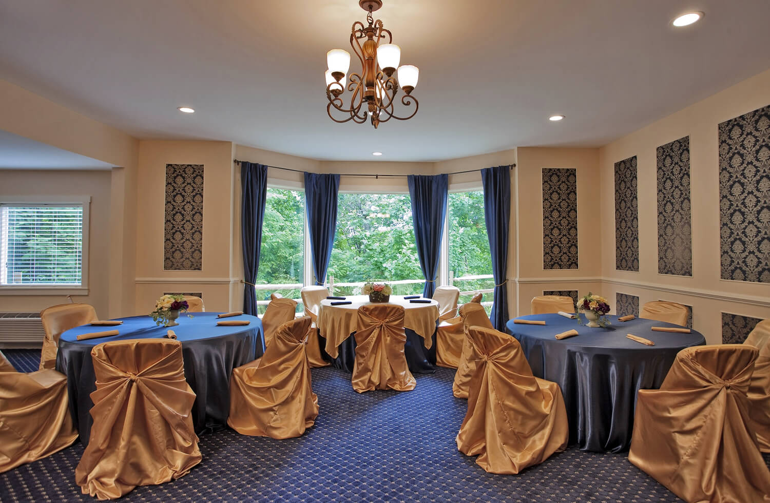 light shines through the windows in the event space, a chandelier hangs above round tables decorated in blue and gold