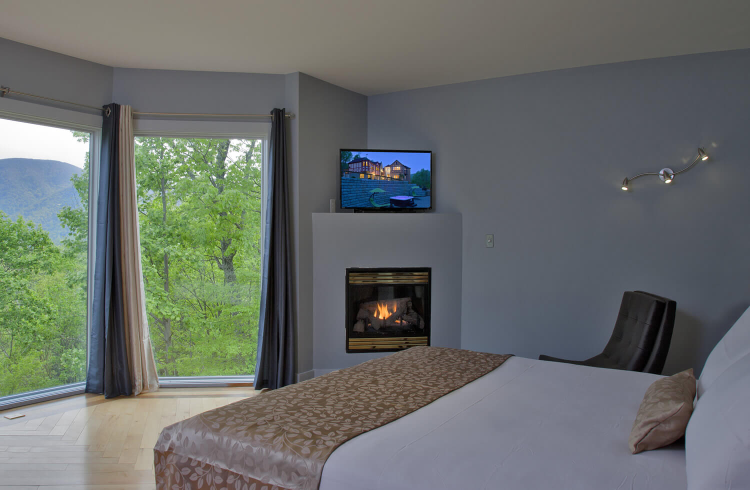 A white bed with gold accents and throw rests across from a fireplace and open window with mountain view